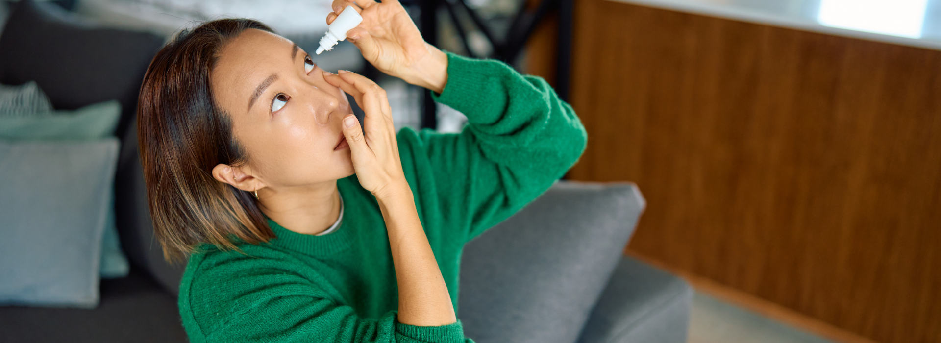 Young adult woman applying eye drops to her open eye while sitting on a couch at home