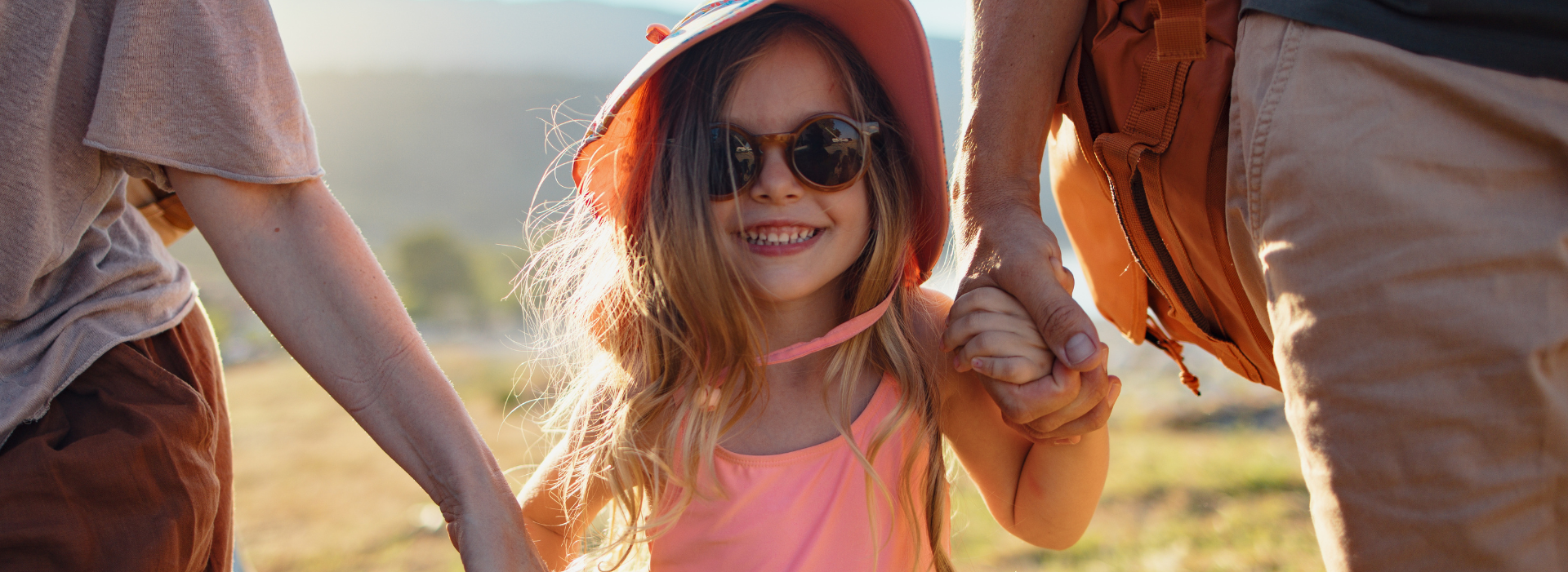 A cheerful young girl wearing a sunhat and sunglasses holding hands with her parents.